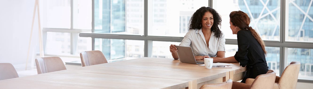 Two young women sitting at the end of a table, looking at a laptop.